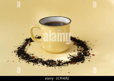 A cup of black tea in a ceramic yellow cup with dry tea scattered on a yellow background. Copy space. Stock Photo