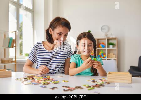 Mom and little daughter put together puzzles while sitting at a table in the room. Stock Photo
