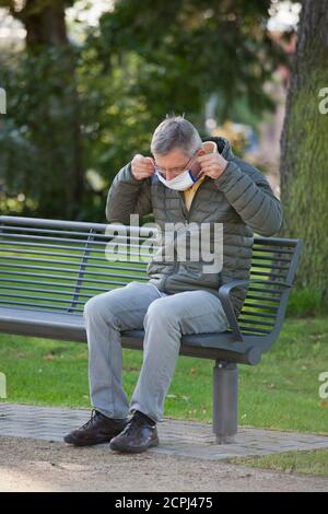 Mature man puts a face mask on or off while sitting on a bech in a park - selective focus on the head Stock Photo