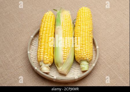 Raw corn cobs in a woven bamboo basket. Isolated on jute background. Close-up. Horizontal shot. Stock Photo