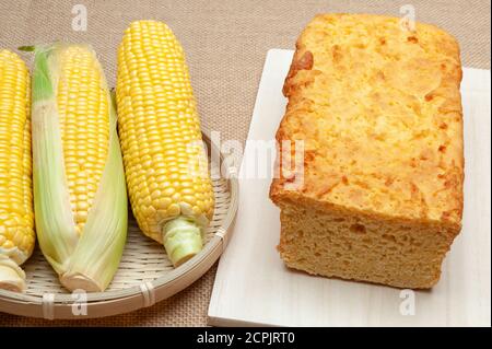 Raw corn cobs in a woven bamboo basket and homemade Brazilian corn bread on a cutting board. Isolated on jute background. Stock Photo