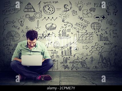 young man working on computer sitting on a floor learning online drawing schemes business plan on a wall Stock Photo