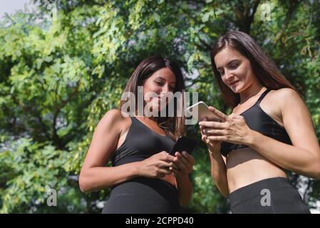 two women dressing black sport clothes checking their smart phones with green trees on the background Stock Photo