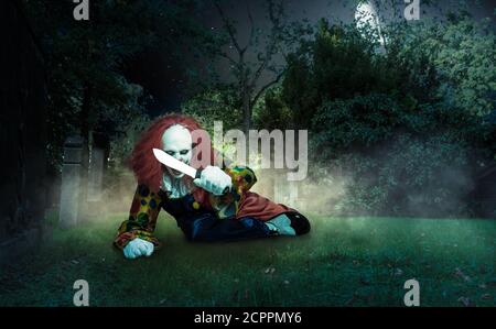 a scary clown lies with a knife on a cemetery threatens the viewer Stock Photo