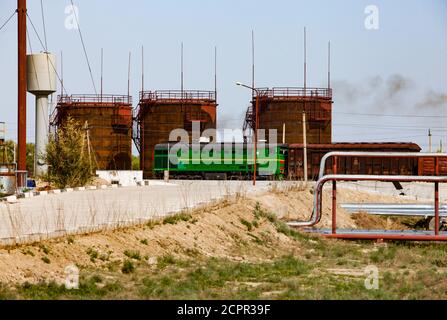 Brown rusted old oil storage tanks under bright sun on blue sky. Railway terminal of refinery plant in desert. Locomotive and carriages. Taraz city, K Stock Photo