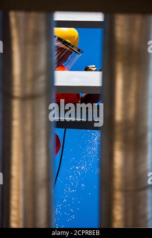 Worker in yellow helmet with angle grinder, glass protective mask and yellow sparks between metal pipes on blue sky background. Stock Photo