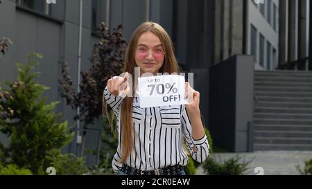 Amazed smiling girl showing Up To 70 percent Off inscriptions signs, pointing at camera, rejoicing good discounts, low prices for online shopping sales. Black Friday concept. Mall on background Stock Photo