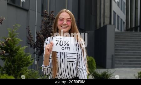 Amazed smiling girl showing Up To 70 percent Off inscriptions signs, rejoicing good discounts, low prices for online shopping sales. Black Friday concept. Supermarket, mall on background Stock Photo