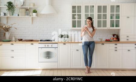 Positive young lady using cell phone in the kitchen Stock Photo