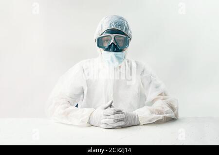 Man in white hazmat on white background studio. Person in a protective suit mask and gloves sits at a table and looks at the camera.  Stock Photo