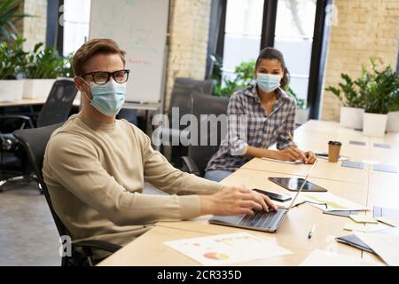 Working safely. Male and female office workers wearing medical protective masks sitting together at the table in the modern office and looking at camera, guy working on laptop. Business and covid19 Stock Photo