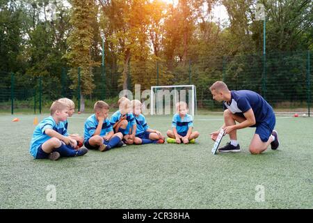 Young coach with clipboard teaches children strategy playing on football field. Stock Photo