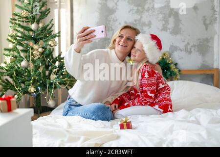 Christmas online family congratulations. Smiling european blonde mother and daughter making selfie photo in Christmas costumes Stock Photo