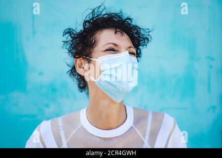 Close up of mature woman with curly short hair wearing safety mask against blue wall Stock Photo