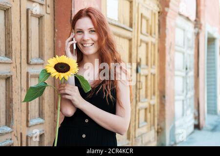 Smiling ginger girl speak on phone at old wooden door background. Woman with a sunflower in hand. Stock Photo