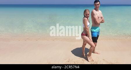 Travel and rest. Young adult man and woman pose against a backdrop of pure blue sea standing on sand, banner Stock Photo