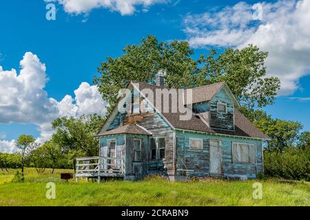 Old, abandoned blue prairie farmhouse with trees, grass and blue sky in Kayville, Saskatchewan, Canada Stock Photo