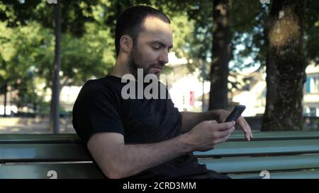 Young handsome man sitting on bench browsing internet via phone. Stock Photo