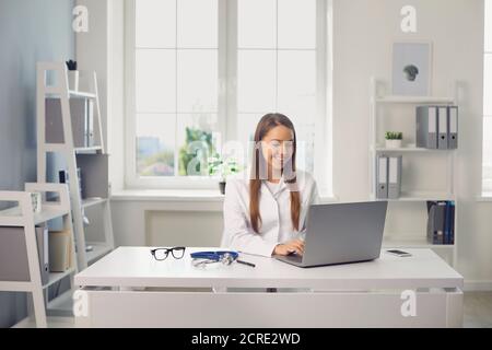 Smiling woman doctor sitting in medical clinic office with laptop and communicating online with somebody Stock Photo
