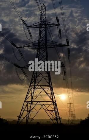 Electric pylon transmission tower silhouette with moody sunrise and electric cables leading to pylons in the distance across agricultural  landscape. Stock Photo