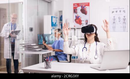 Young medic doing hand gesture wearing virtual reality headset in hospital office. Doctor in white coat with stethoscop. Senior physician writing notes on clipboard. Nurse in blue uniform looking at x-ray. Stock Photo