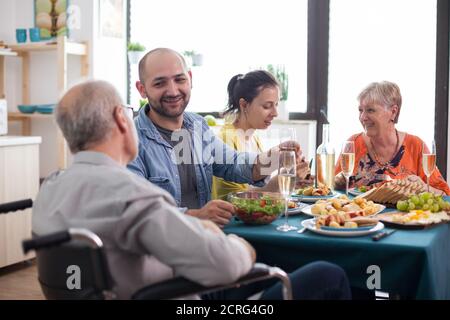 Son smiling at disabled senior father in wheelchair during family gathering. Old mother having a conversation with daughter while eating lunch in kitchen. Stock Photo