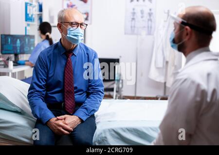 Sick patient talking with doctor during consultation wearing protective mask against covid in hospital consultation room. Global health crisis, medical system during pandemic, sick elderly patient. Stock Photo