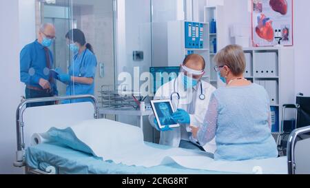 Doctor in protective visor talking with patient wearing a mask explainingX ray result on digital tablet PC in modern private clinic or hospital. Medical stuff working after coronavirus outbreak Stock Photo
