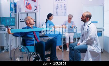 Senior man in wheelchair exercising muscle strength with elastic band under medical supervision in modern recovery clinic or hospital. Invalid physiotherapy program, health care injury rehabilitation Stock Photo