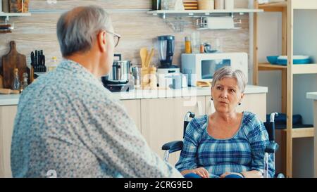 Handicapped senior woman in wheelchair bonding with husband while food is getting ready on gas cooker. Old man having a conversation with wife. Living with disabled person with walking disabilities Stock Photo
