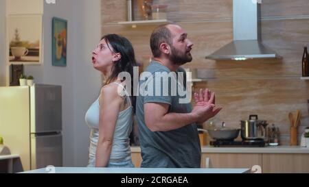 Couple with trust issues fighting back to back. Furious, irritated, frustrated, jealous unhappy couple screaming accusing to each other having family conflict disputing sitting in the kitchen. Stock Photo