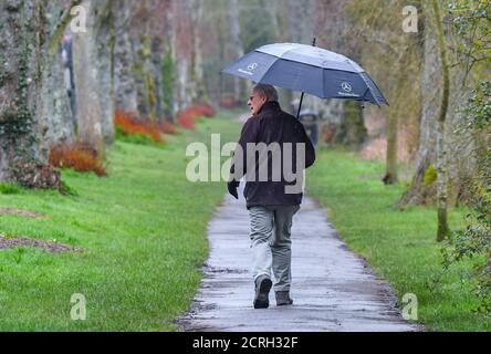 Senior elderly man walking in the rain along a path holding an umbrella while it's raining in England, UK. Stock Photo