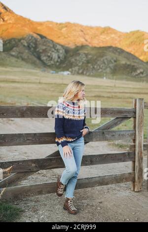 Young blonde woman in nordic sweater and jeans near old wooden fence on mountains background Stock Photo