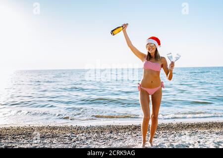 Beautiful blond female in red and white bikini and Santa hat with champagne on a beach. Christmas, new year in tropics. Vacation, holidays Stock Photo