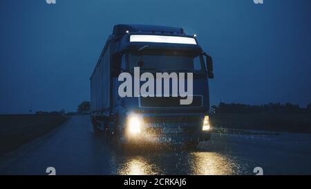 Blue Long Haul Semi-Truck with Cargo Trailer Full of Goods Travels At Night on the Freeway Road, Driving Across Continent Through Rain, Fog, Snow Stock Photo