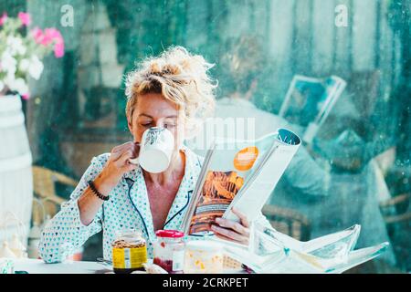 Blonde young mature woman in pyjamas at home in breakfast time, reading a magazine and having a cup of coffee Stock Photo