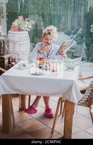 Blonde young mature woman in pyjamas at home in breakfast time, reading a magazine and eating a toast. Stock Photo