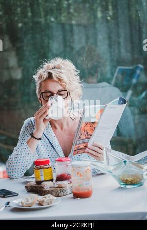 Blonde young mature woman with glasses in pyjamas at home in breakfast time, reading a magazine and having a cup of coffee Stock Photo