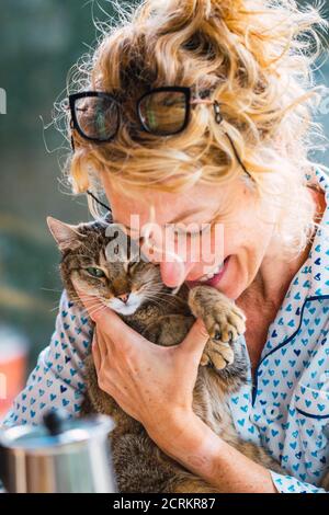 Blonde young mature woman with glasses in pyjamas at home in breakfast time, reading a magazine and having a moment with his cat. Stock Photo