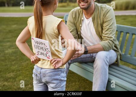 Father sitting on the wooden bench in park while his little daughter hiding handmade postcard for him, small girl making a surprise for Happy Fathers Stock Photo