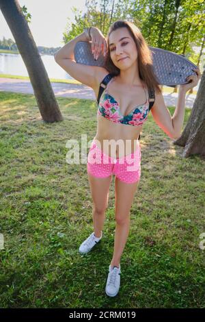 sports young woman in bikini at sunny park standing on skateboard, looking at camera, smiling Stock Photo