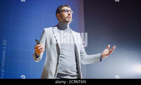 On-Stage Handsome Speaker Greets Audience, Talks about His Ideas, Concepts and New Age Product, in Background Screen Animation. Live Entrepreneurship Stock Photo