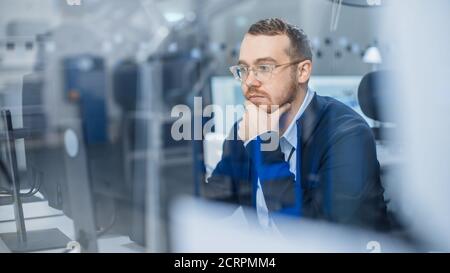 Shot of Thoughtful Industrial Engineer Working on a Personal Computer. He is Working in Cozy Office in Modern Factory with High-Tech Machinery. Stock Photo