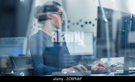 Shot Through Glass. Female Engineer Working on Personal Computer. She is Working in Office in Modern Factory with High-Tech Machinery. Stock Photo