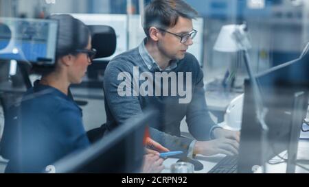 Shot Through Glass in Modern Factory: Female Project Supervisor Talks to a Male Industrial Engineer who Works on Personal Computer. Modern High-Tech Stock Photo