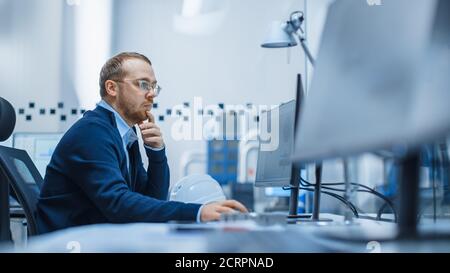 Shot of Male Industrial Engineer Solving Problems, Working on a Personal Computer, He is Working in Office in Modern Factory with High-Tech Machinery. Stock Photo