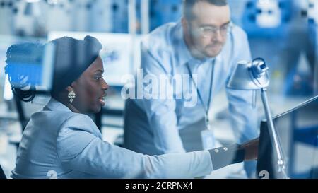 Modern Factory: Male Project Supervisor Talks to a Female Industrial Engineer who Works on Personal Computer. Shot Through Glass in Modern High-Tech Stock Photo