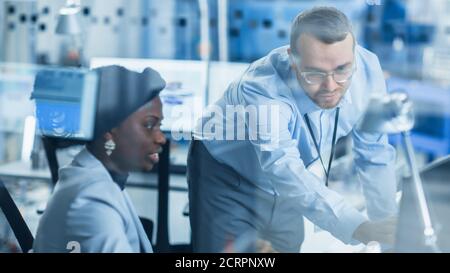 Modern Factory: Male Project Supervisor Talks to a Female Industrial Engineer who Works on Personal Computer. Shot Through Glass in Modern High-Tech Stock Photo