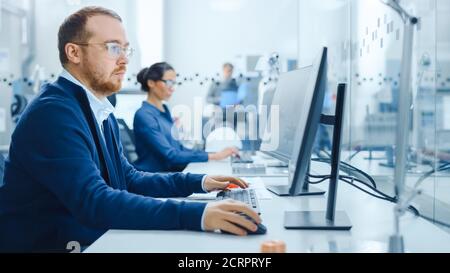 Inside Factory Glass Office Team of Industrial Engineers Working on PC, Outside Cubicle in the Modern Facility Workshop Professional Workers Using Stock Photo