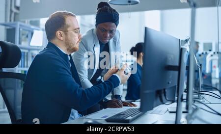 Modern Factory: Female Project Supervisor Talks to a Male Industrial Engineer who Works on Computer. They use CAD Software to Design Industrial Stock Photo